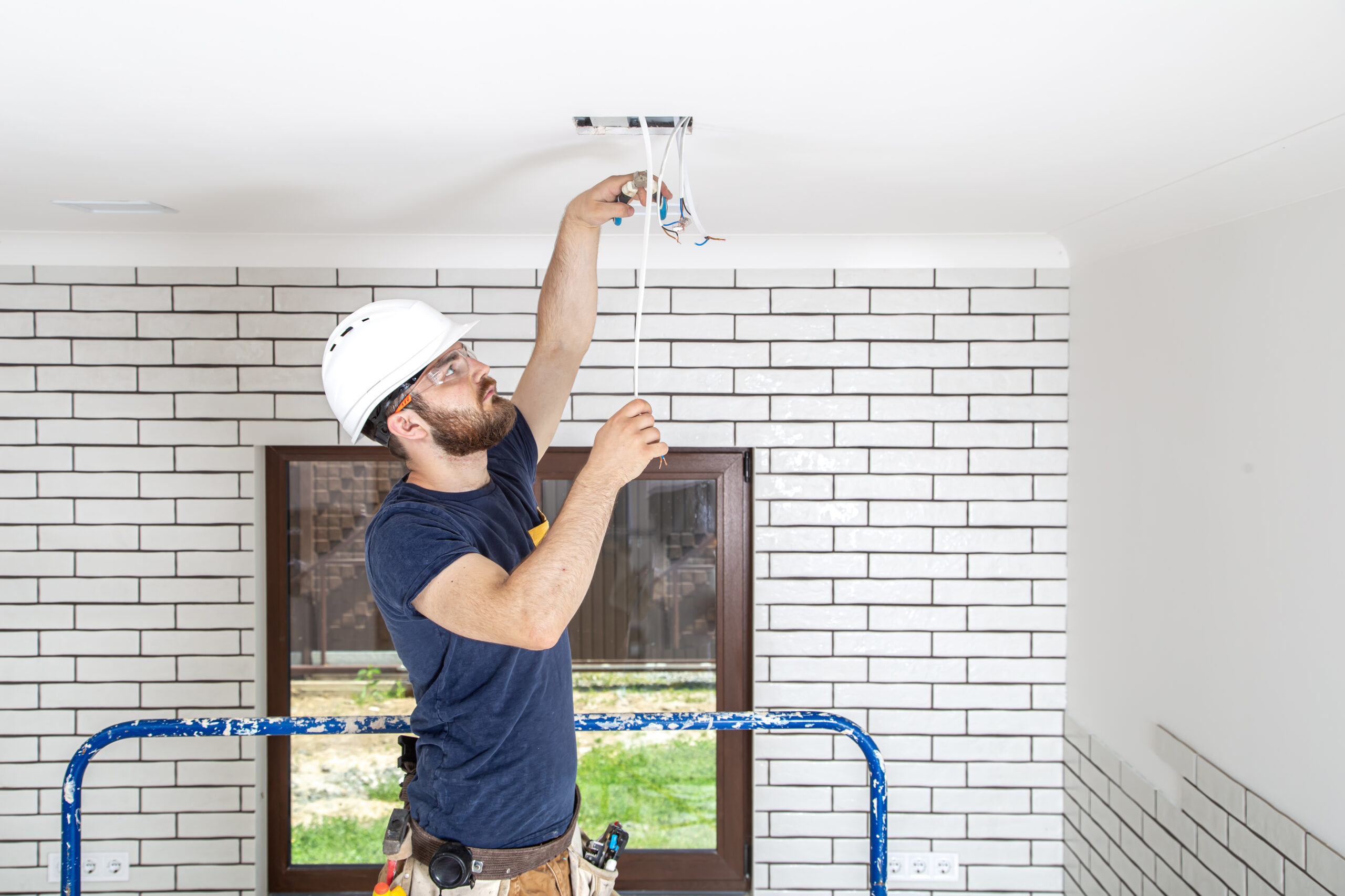 Electrician Builder at work, installation of lamps at height. Professional in overalls with an electrical tool. On the background of the repair site.
